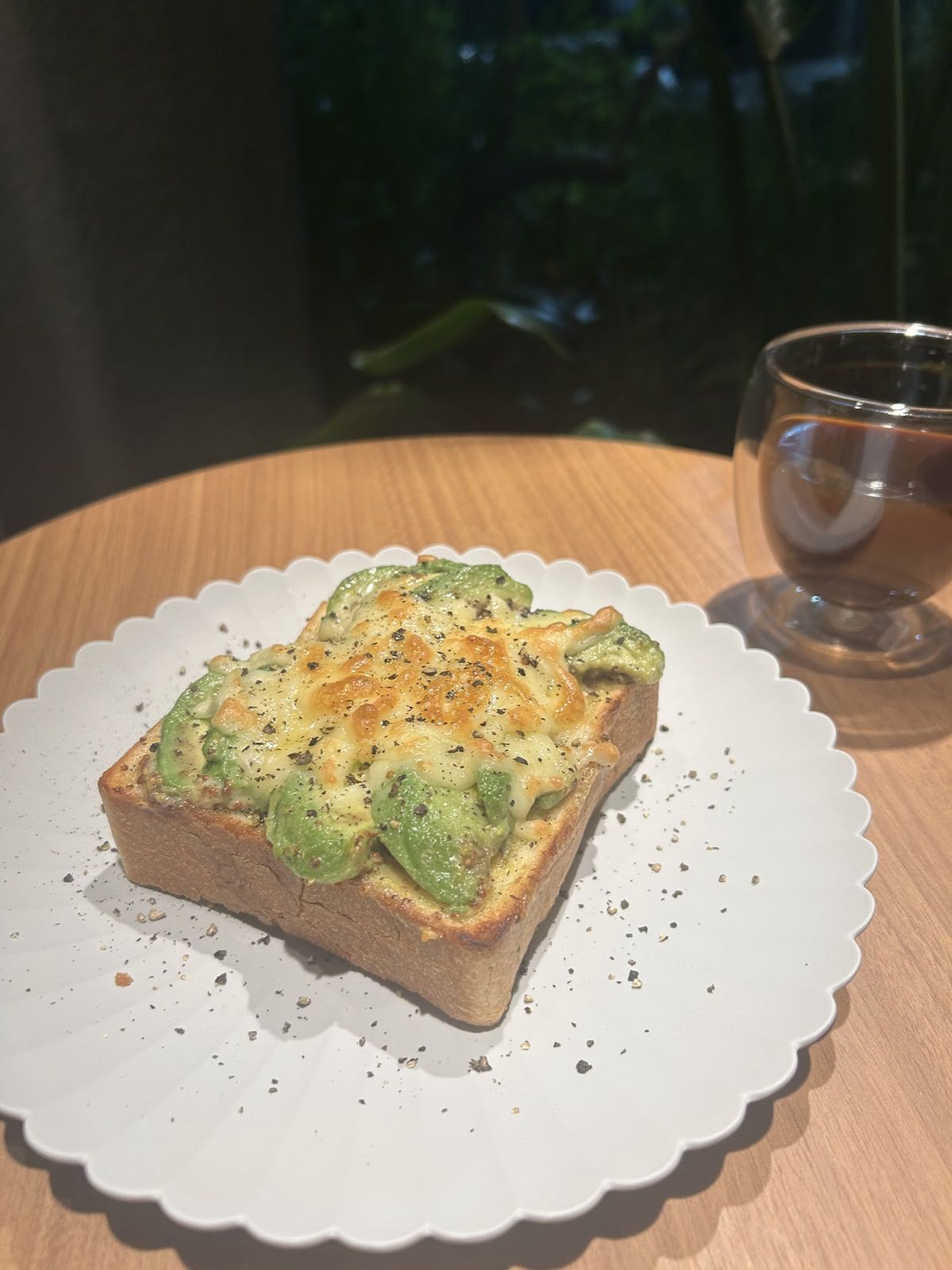 白い皿にのったトースト。アボカドとチーズを焼いて黒胡椒をふってあり、横にガラスのカップのコーヒー。窓の外は緑。