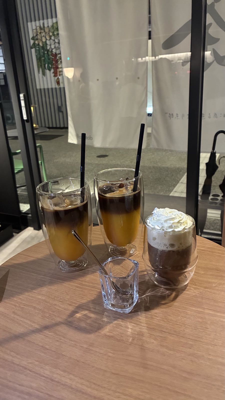 木のテーブルに並んだ三杯。二重ガラスのグラスに入ったコーヒーと果汁の二層、右にクリームを乗せた一杯。奥に白い暖簾。